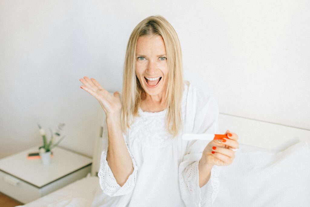 A joyful woman celebrates a positive pregnancy test while sitting on her bed.